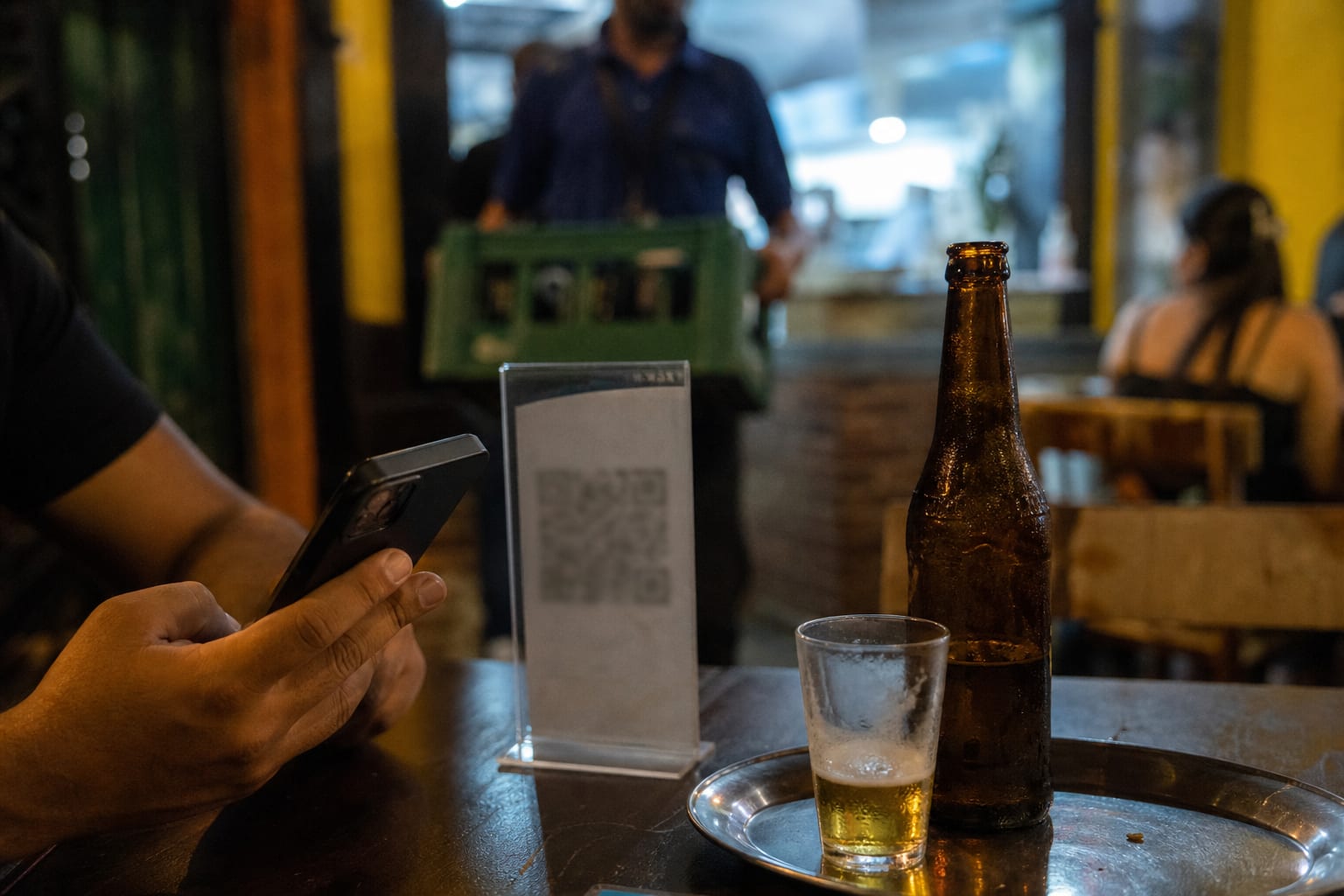 Cliente lendo o QR de um display de acrílico na mesa enquanto o garçom passa ao fundo